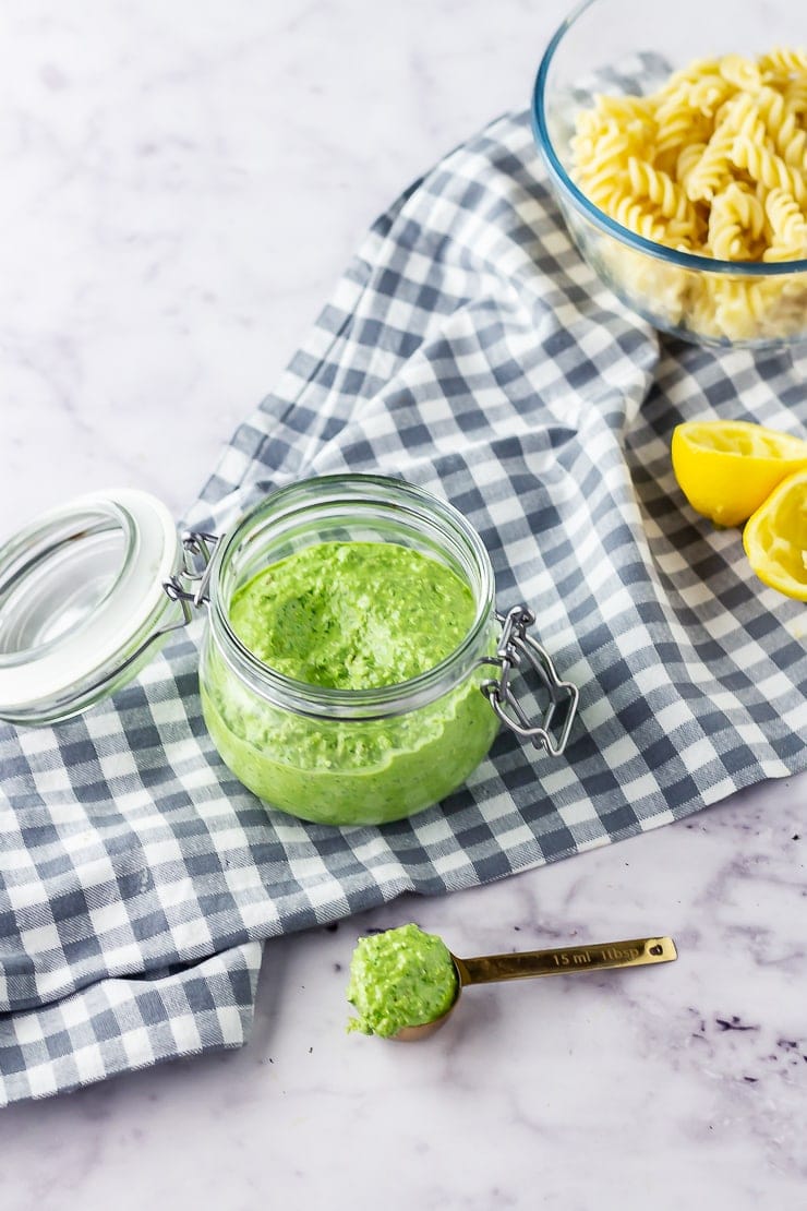 Jar of spinach pesto on a checked cloth with lemon and pasta in the background