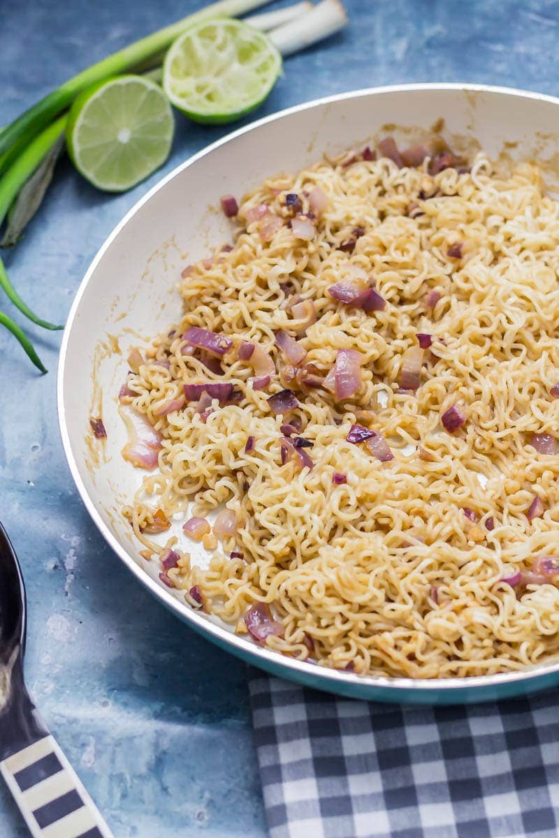 Frying pan of soy peanut noodles on a blue surface with limes and spring onions