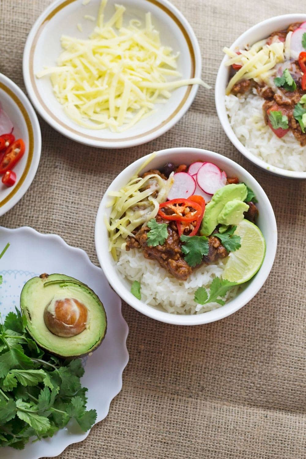 Overhead shot of bowls of classic beef chilli con carne with grated cheese and avocado on a hessian background