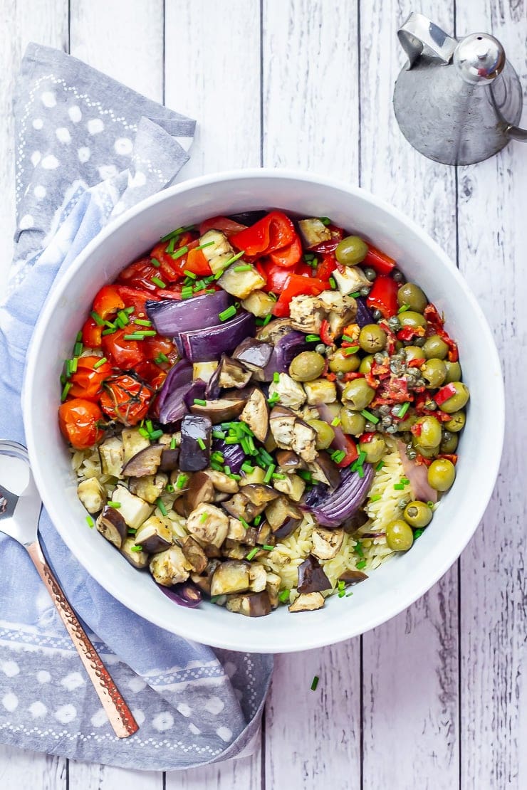 Overhead shot of summer orzo salad on a white wooden background with cloth, spoon and olive oil jug