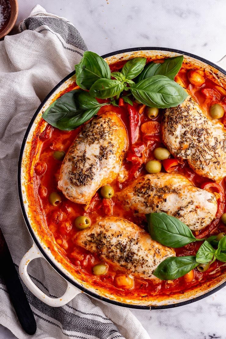 Overhead shot of white pot of chicken in tomato sauce with basil on a marble background