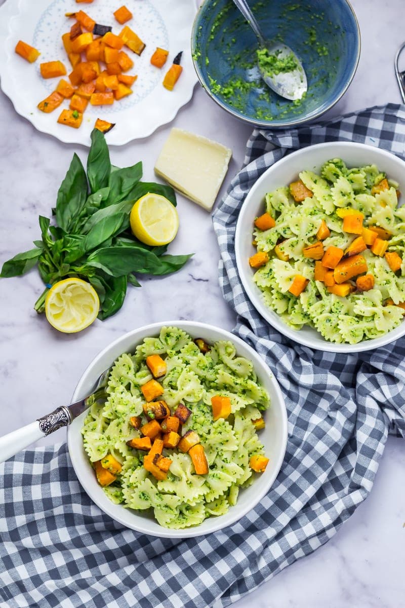 Two bowls of kale pesto bowties and roasted butternut squash with lemon, basil leaves and more butternut squash on a marble background with a checked cloth