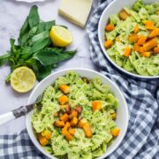 Two bowls of kale pesto pasta with a checked cloth underneath and pesto ingredients in the background