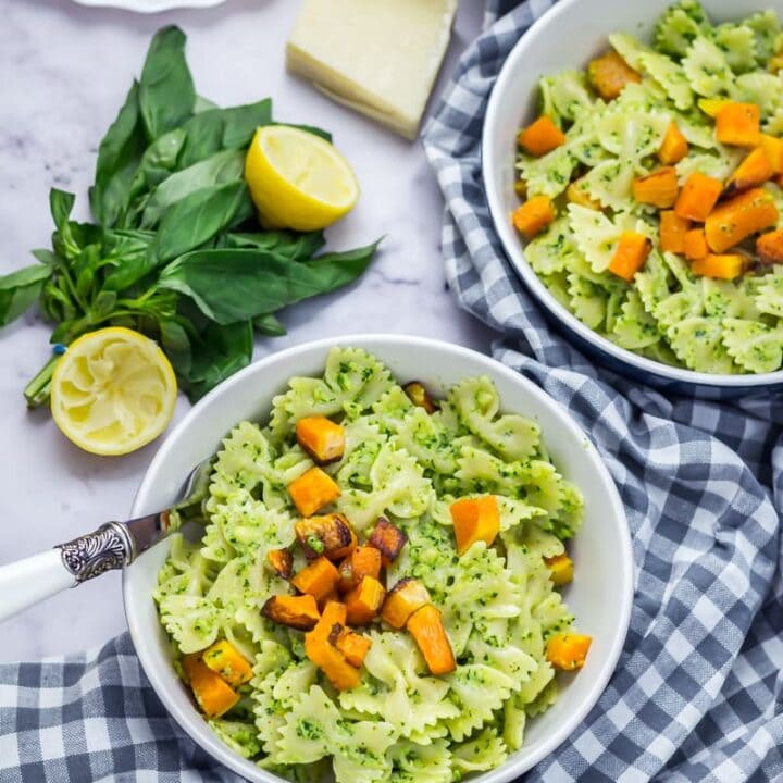 Two bowls of kale pesto pasta with a checked cloth underneath and pesto ingredients in the background