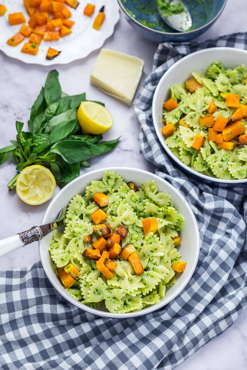 Two bowls of kale pesto pasta with a checked cloth underneath and pesto ingredients in the background