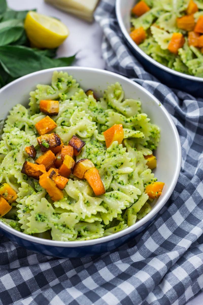 Bowl of kale pesto pasta on a checked cloth with another bowl and lemon and basil leaves in the background