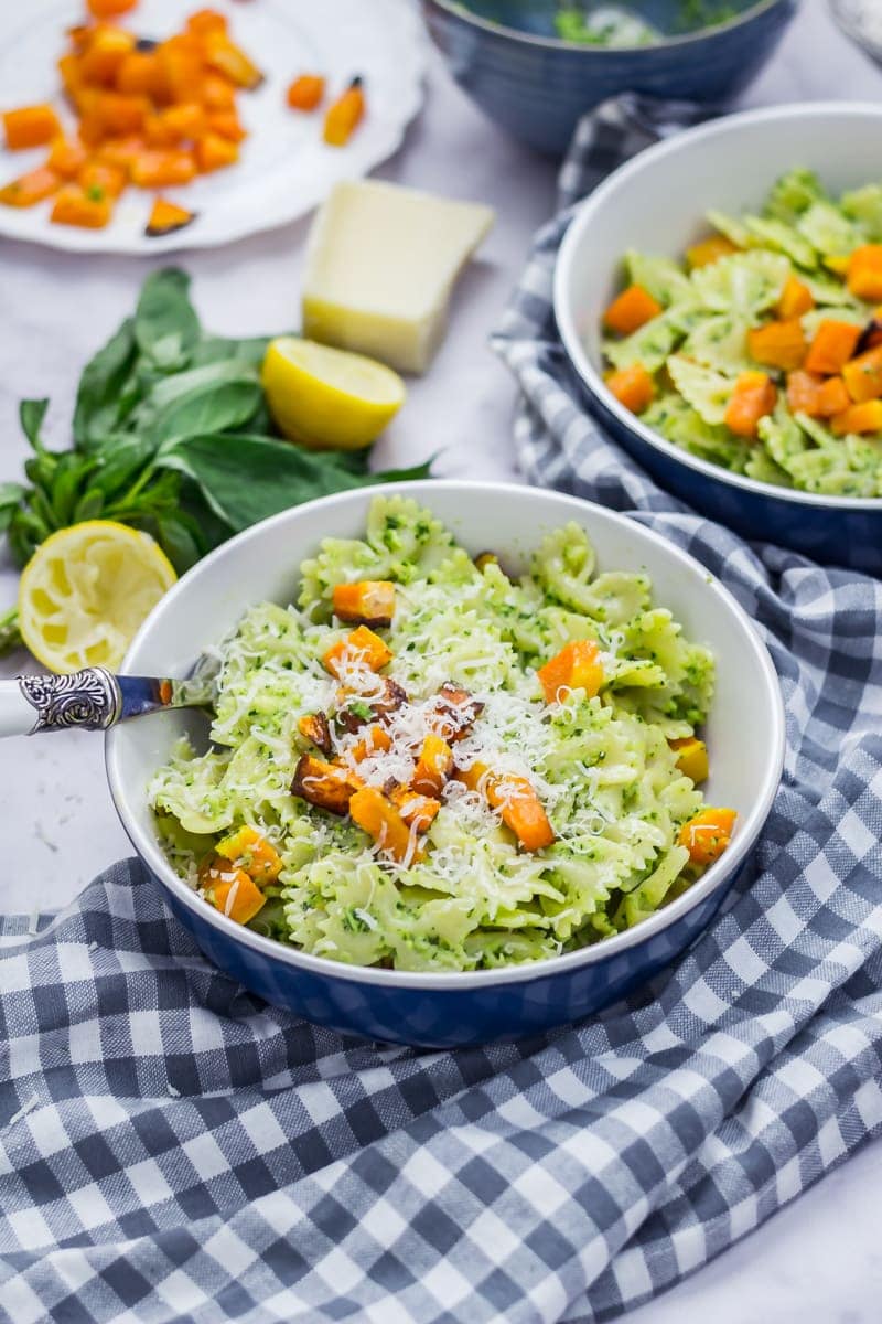 Two bowls of kale pesto pasta with butternut squash on a checked cloth with basil, lemon halves, parmesan and butternut squash cubes in the background