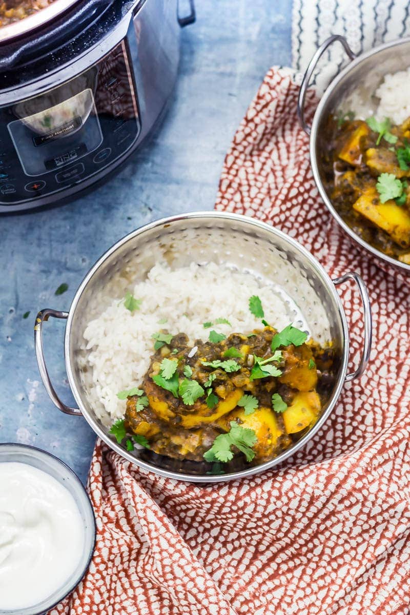 Two bowls of saag aloo with rice topped with coriander leavers next to a bowl of yoghurt and a pressure cooker on a blue background