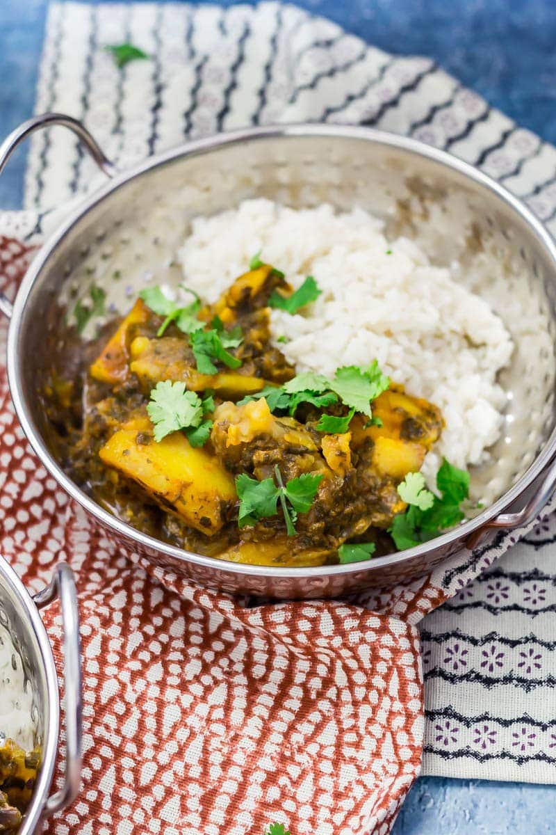 Metal bowl of saag aloo with rice, topped with coriander leaves on a patterned tea towel