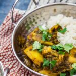 Metal bowl of saag aloo with white rice topped with coriander leaves on a patterned tea towel