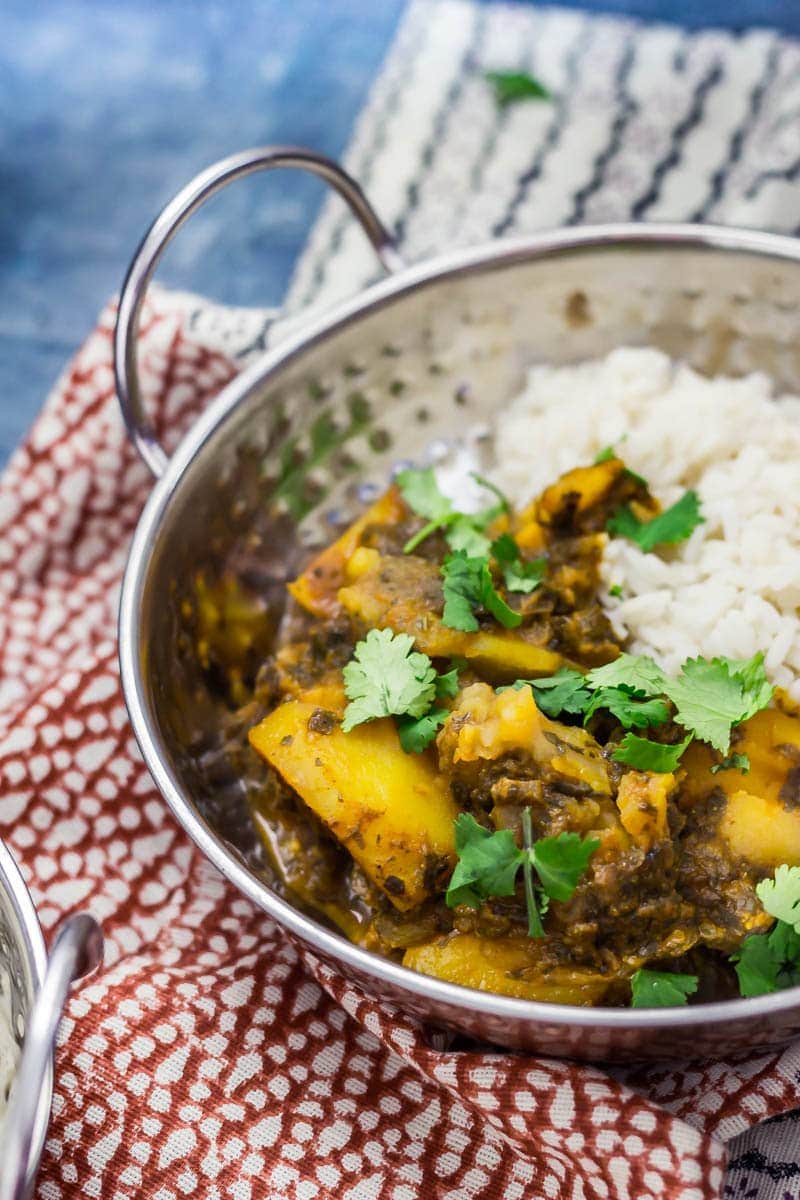 Metal bowl of saag aloo with white rice topped with coriander leaves on a patterned tea towel