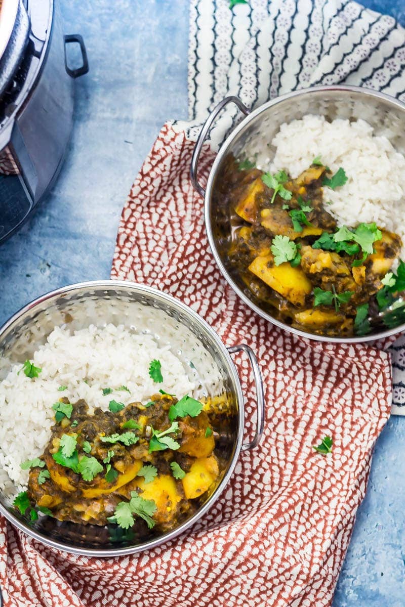 Two metal bowls of saag aloo with rice and coriander leaves on a patterned towel with a pressure cooker to the side