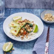 Udon Noodle Stir Fry topped with pak choi on a white bowl with a striped napkin, chopsticks and a bowl of peanuts on the side