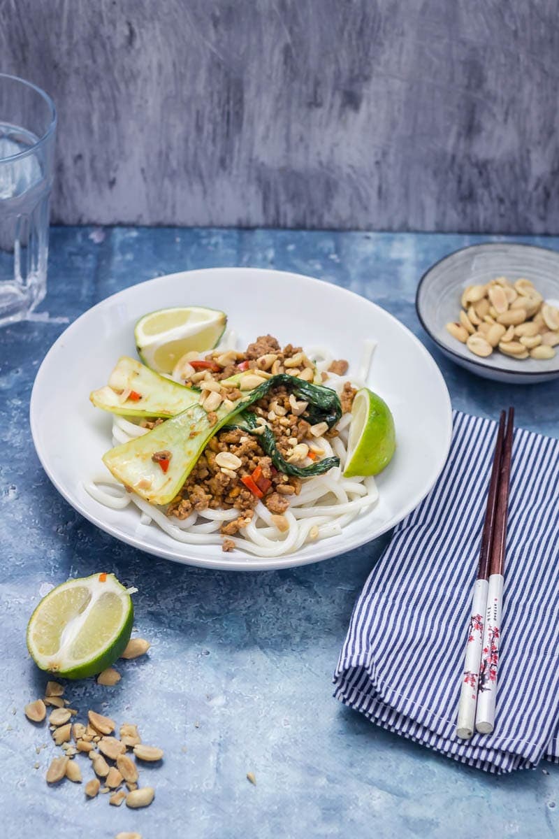 Udon Noodle Stir Fry topped with pak choi on a white bowl with a striped napkin, chopsticks and a bowl of peanuts on the side