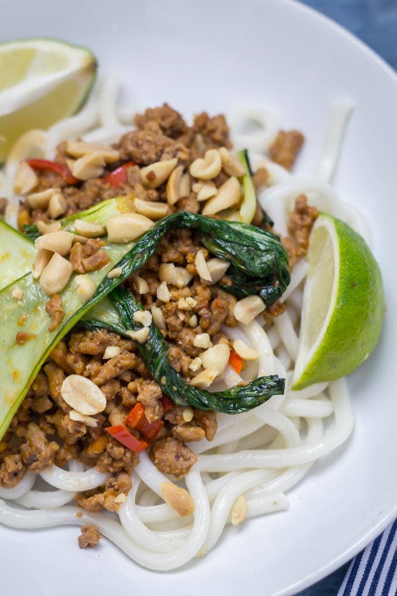 Close up of udon noodle stir fry with turkey mince, pak choi, chilli, peanuts and lime wedges on a white plate