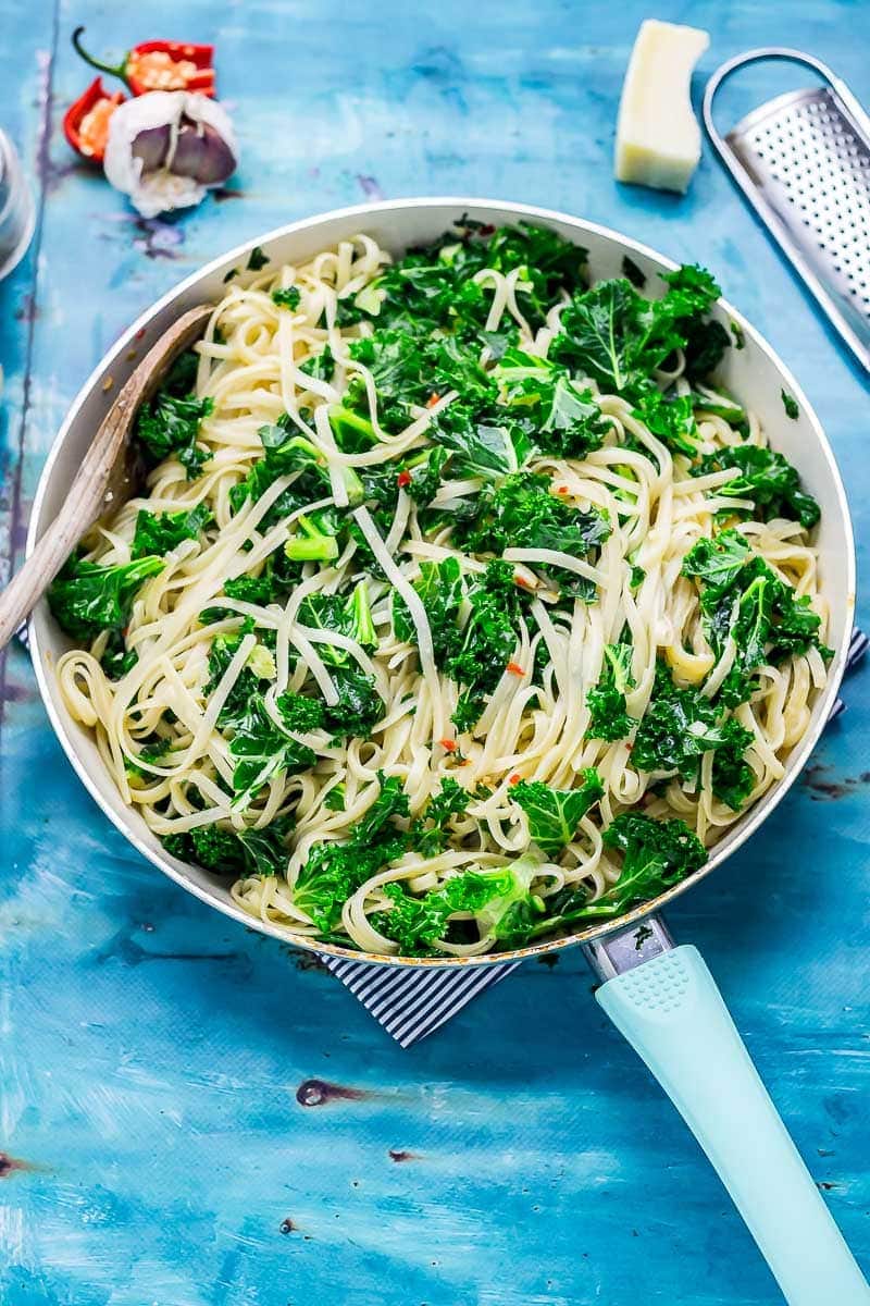 Frying pan of kale pasta on a blue background with parmesan and grater