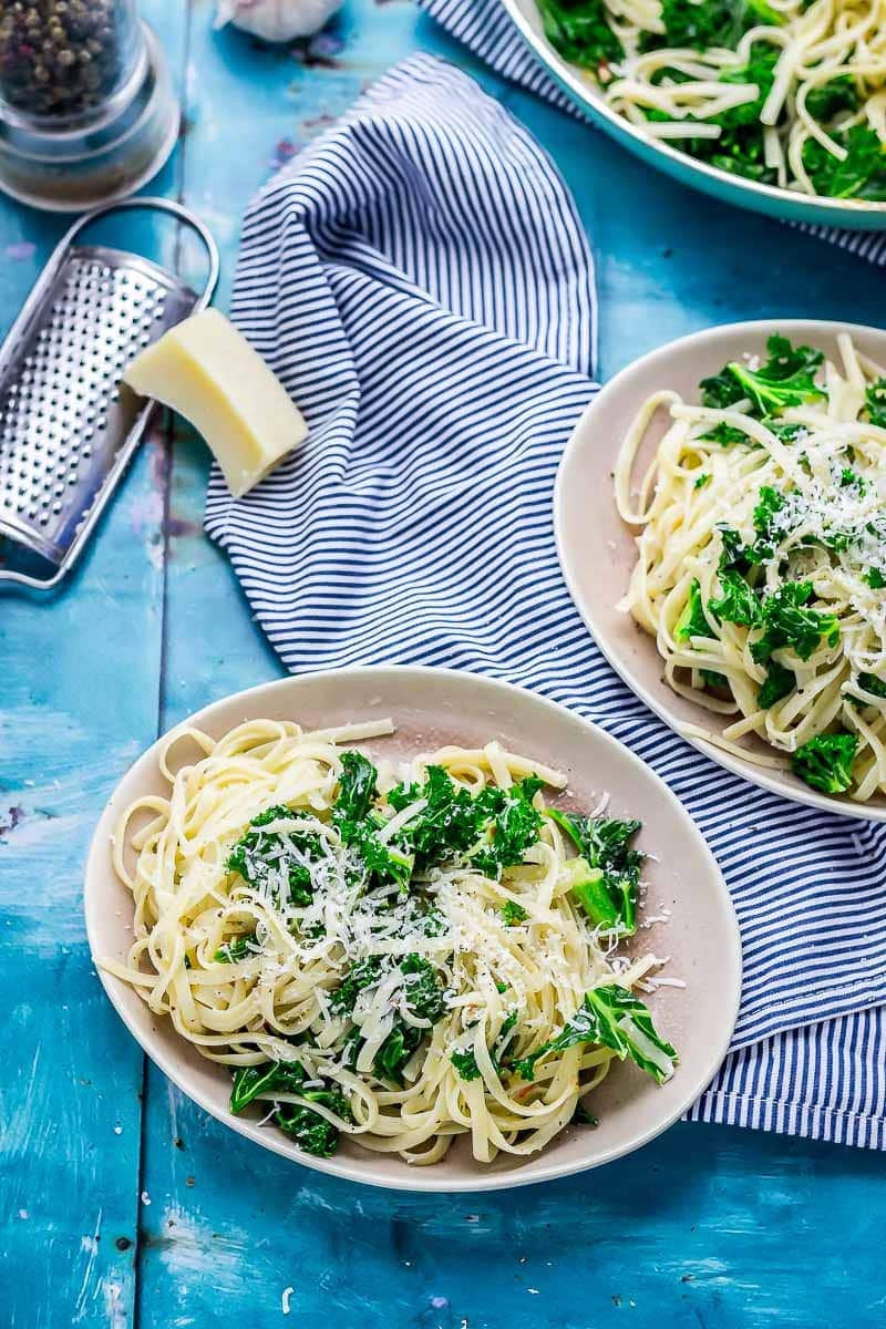 Two plates of kale pasta on a striped cloth with parmesan and a grater on a blue background