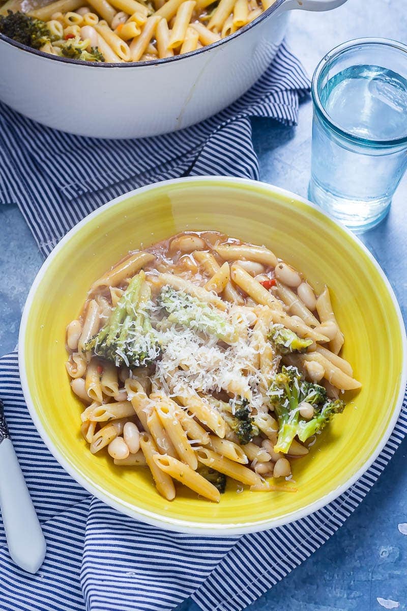 Yellow bowl of Broccoli and White Bean One Pot Pasta on a striped cloth with a glass of water