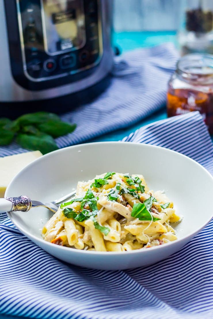 Bowl of creamy chicken pasta on a striped cloth with a pressure king pro and sun dried tomatoes in the background