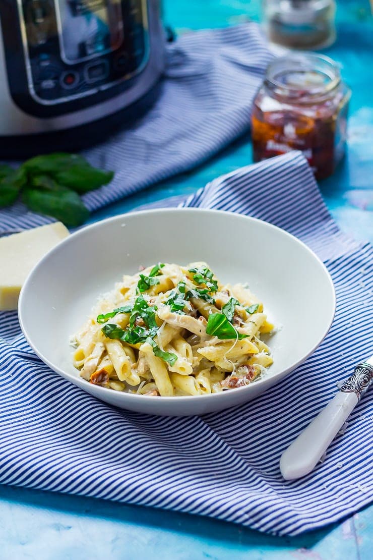 Bowl of creamy chicken pasta on a striped cloth with sun dried tomatoes, parmesan and pressure cooker in the background