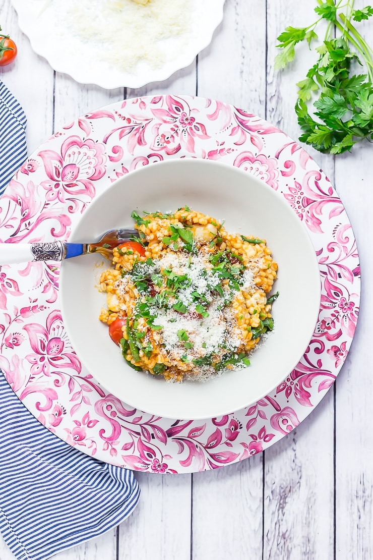 Overhead shot of One Pot Cheesy Pearl Barley with Tomatoes in a bowl on a pink platter with parsley, parmesan and a striped cloth to the side