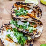 Three chipotle chicken tacos on a wooden board with lime wedges and sour cream