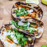 Three chipotle chicken tacos on a wooden board with lime wedges and sour cream