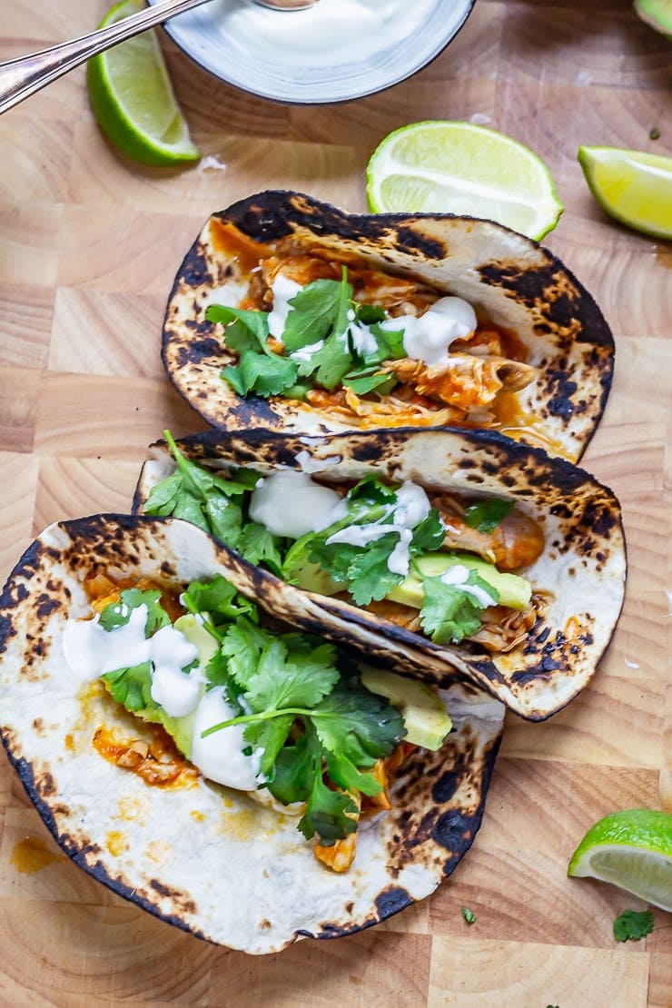 Three chipotle chicken tacos on a wooden board with lime wedges and sour cream