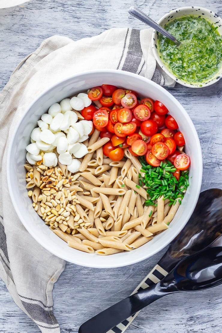 Overhead shot of caprese pasta salad ingredients with cloth, basil dressing and salad servers