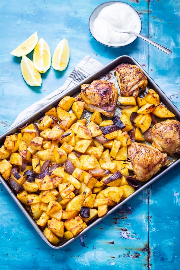 Overhead shot of sheet pan harissa chicken on a blue background with lemon and yoghurt