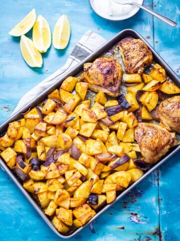 Overhead shot of sheet pan harissa chicken on a blue background with lemon and yoghurt