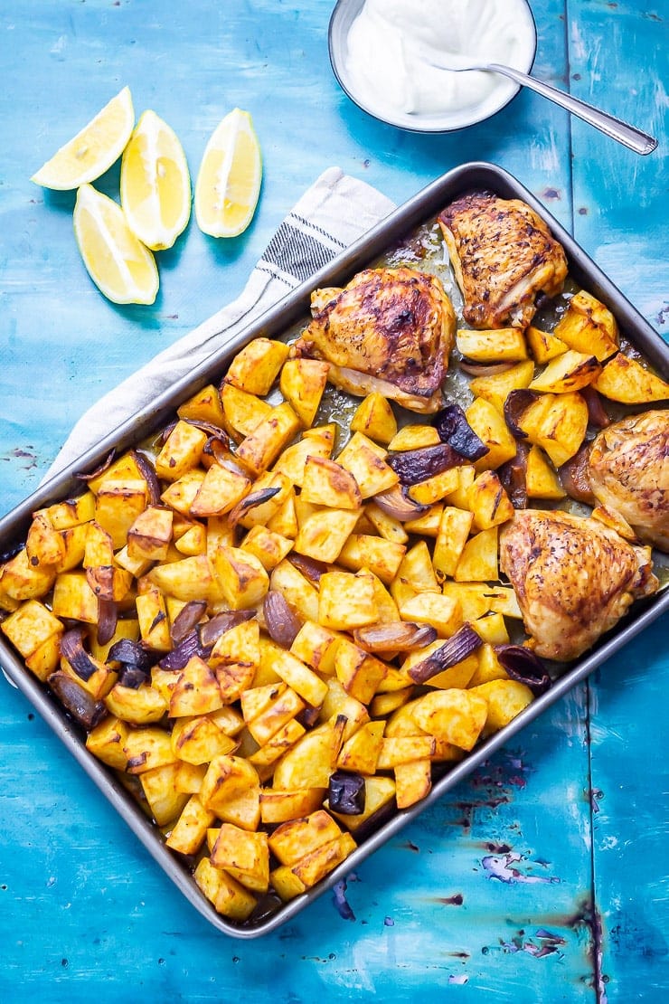 Overhead shot of sheet pan harissa chicken on a blue background with lemon and yoghurt