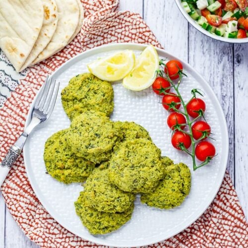 Overhead shot of baked falafel with cherry tomatoes and lemon slices on a patterned cloth and white wooden background