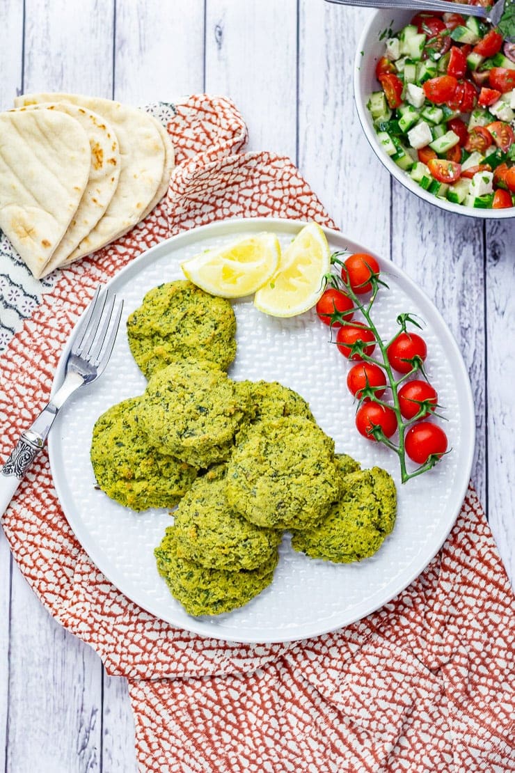 Overhead shot of baked falafel with cherry tomatoes and lemon slices on a patterned cloth and white wooden background