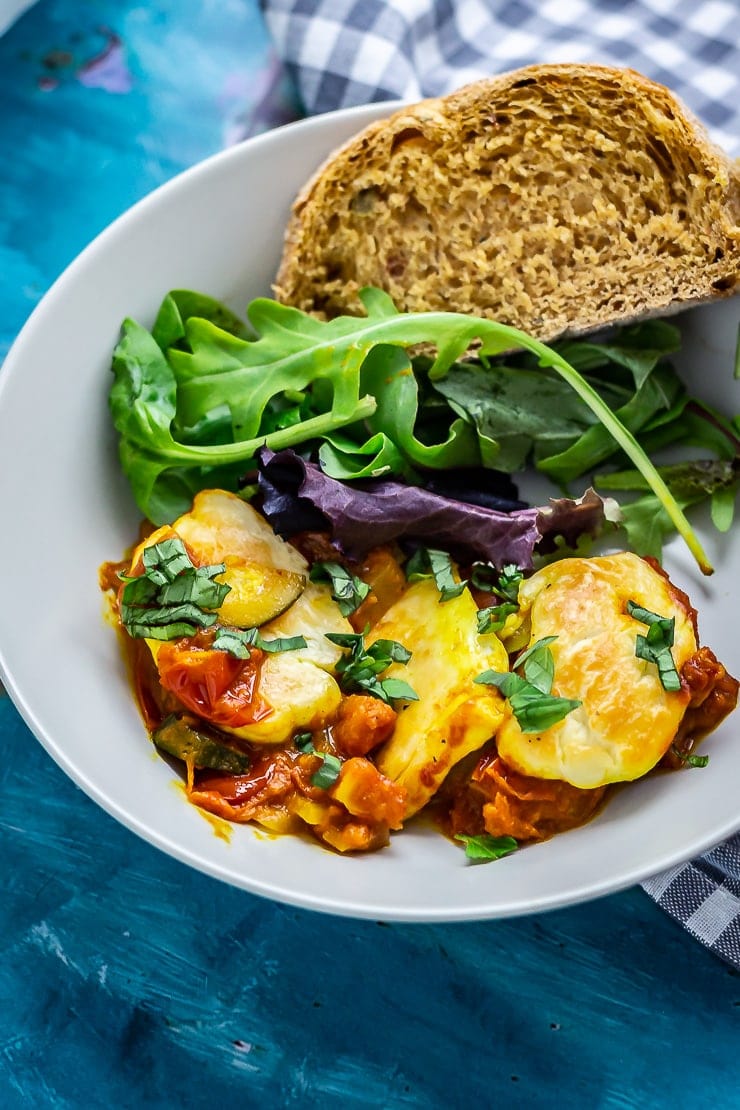 Bowl of tomato and halloumi bake with salad and bread on a blue background