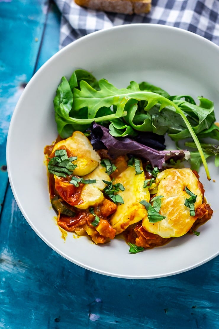 Bowl of tomato and halloumi bake with salad on a blue background
