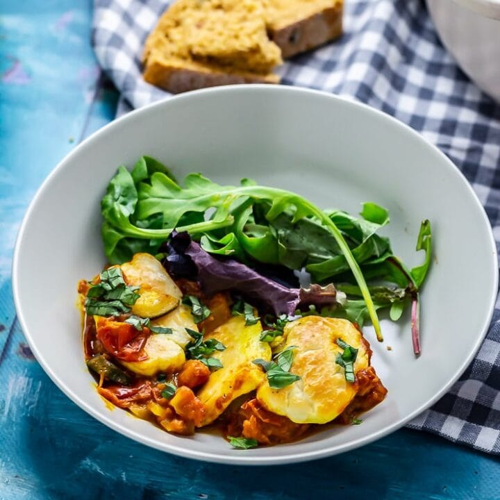 Bowl of tomato and halloumi bake on a blue background with checked cloth in background