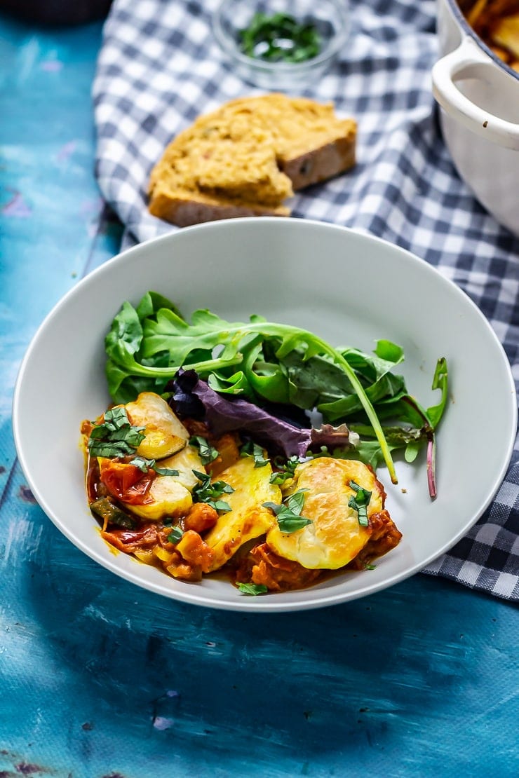 Bowl of tomato and halloumi bake on a blue background with checked cloth in background