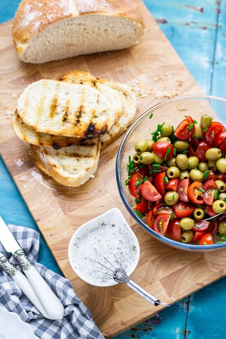 Overhead shot of tomato and olives in a bowl with toast and dressing on a wooden board over a blue background