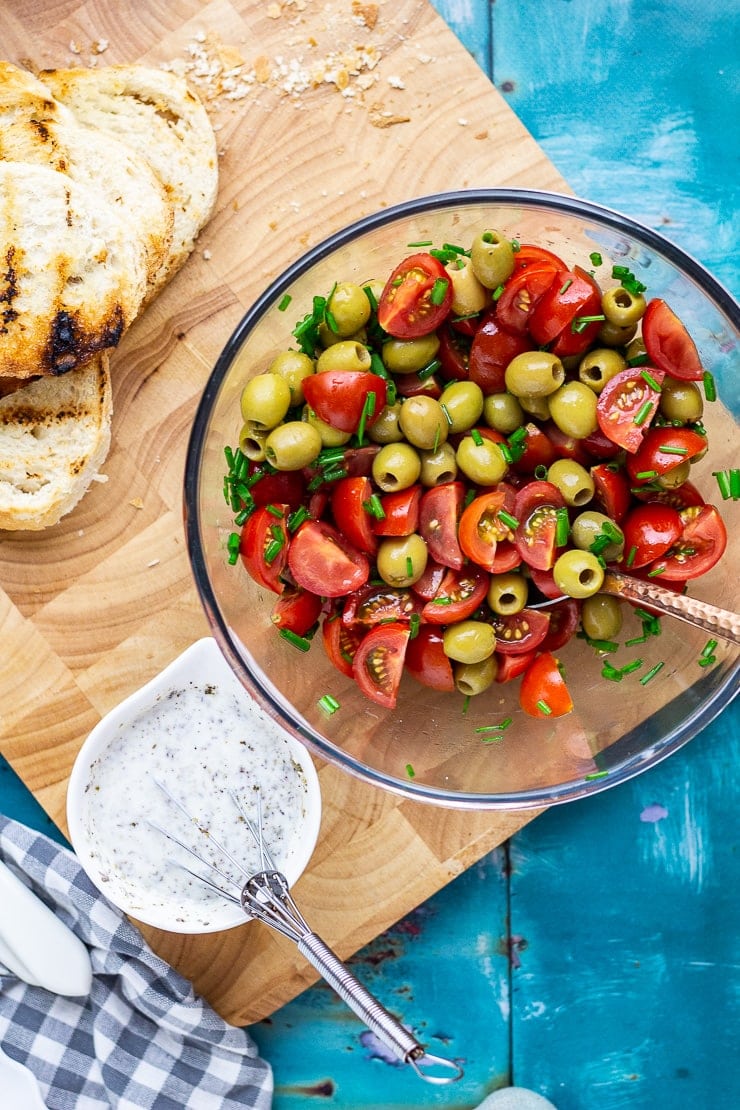 Bowl of olives and tomatoes on a wooden board with dressing and bread on a blue background