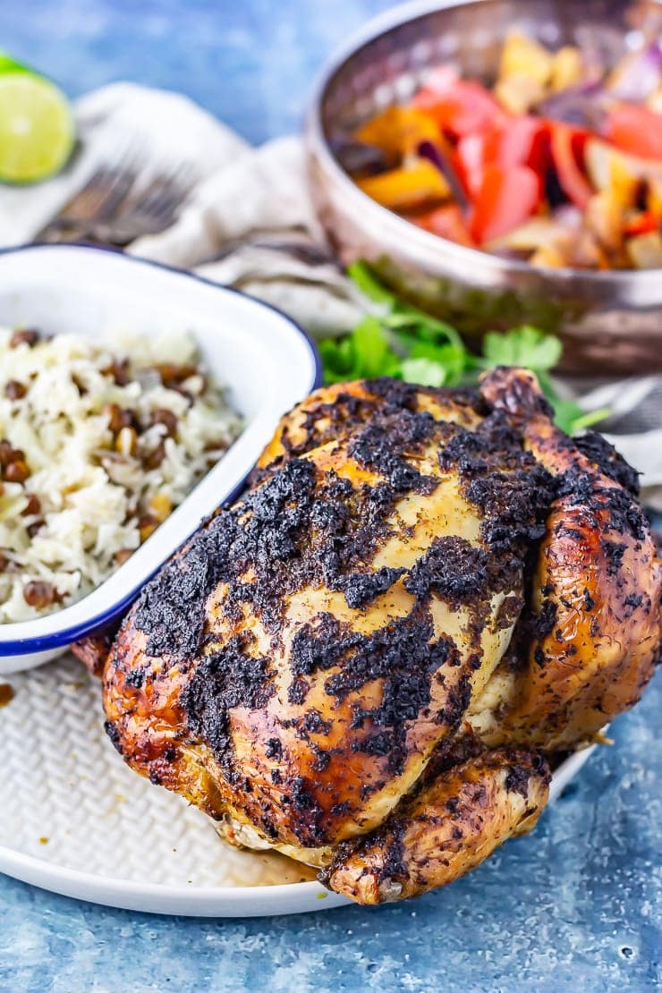 Side shot of jerk roasted chicken on a plate with rice and curry in the background on a blue background