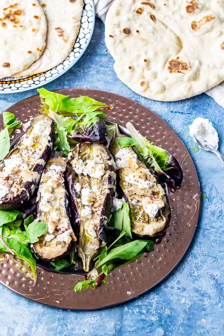 Overhead shot of baked aubergine with tahini on a platter with flatbread on a blue background