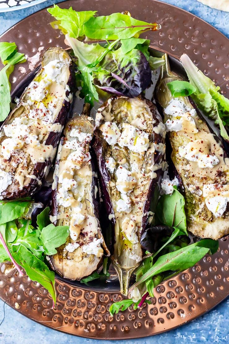 Overhead close up shot of baked aubergine with tahini and goat's cheese on a platter over a blue background