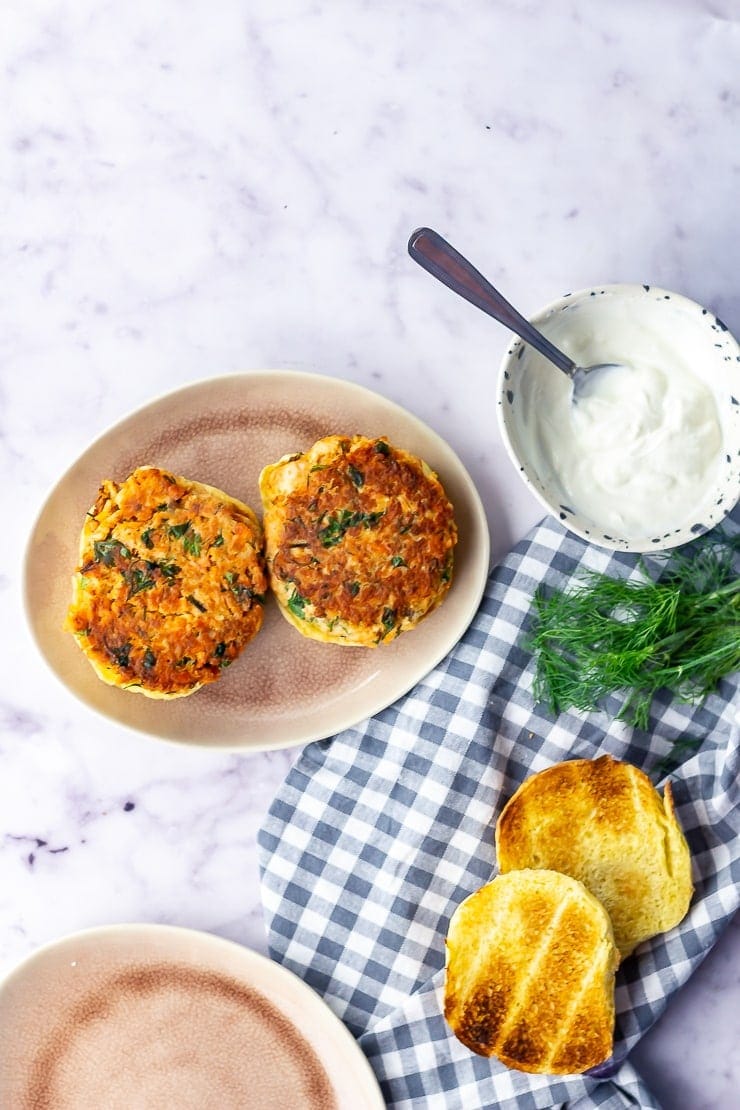 Overhead shot of salmon burgers on a plate with toasted buns on a checked cloth over a marble background