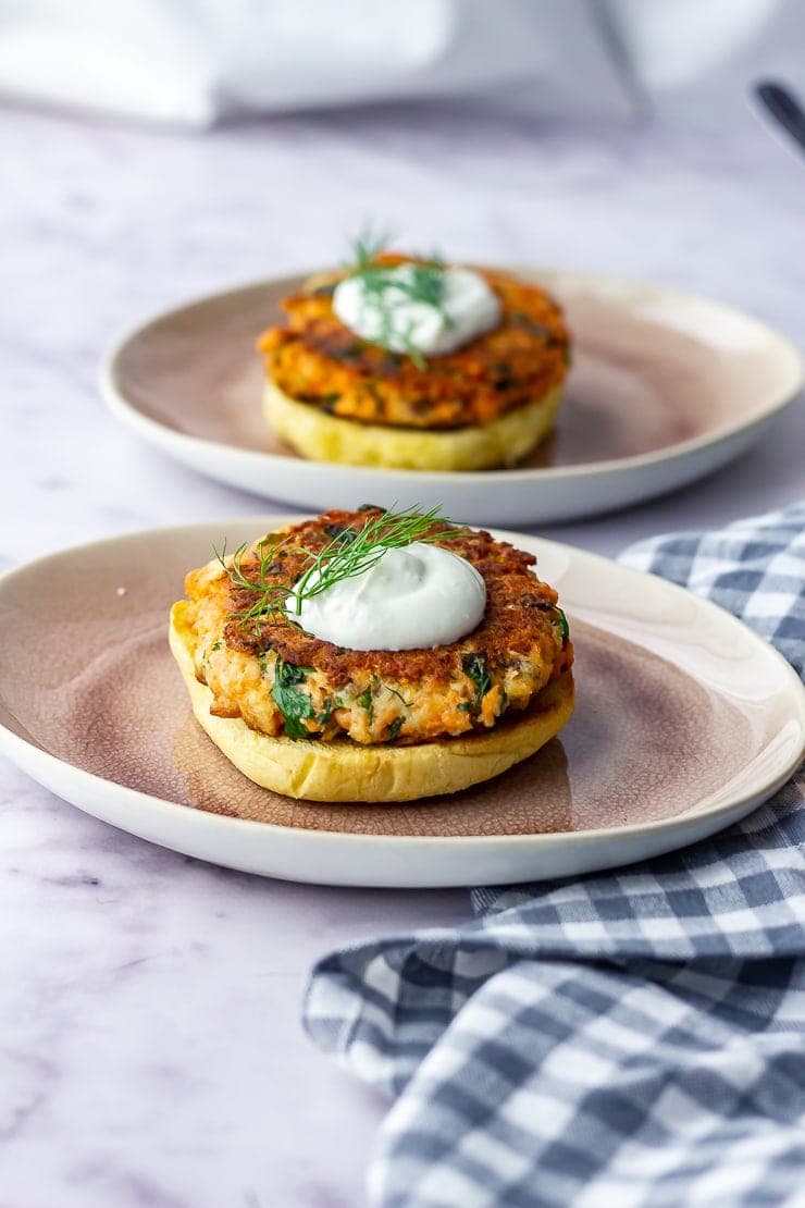 Photo of two salmon burgers on buns on pink plates with a checked cloth on a marble background