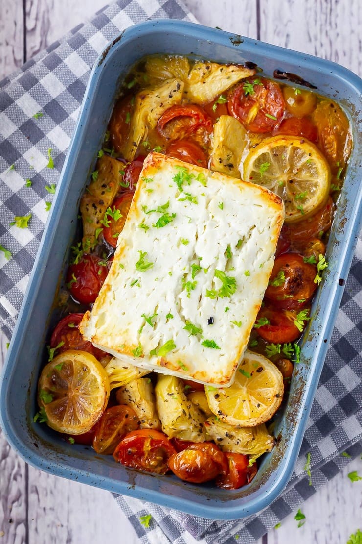 Overhead shot of baked feta with roasted vegetables in a blue baking dish on a checked cloth
