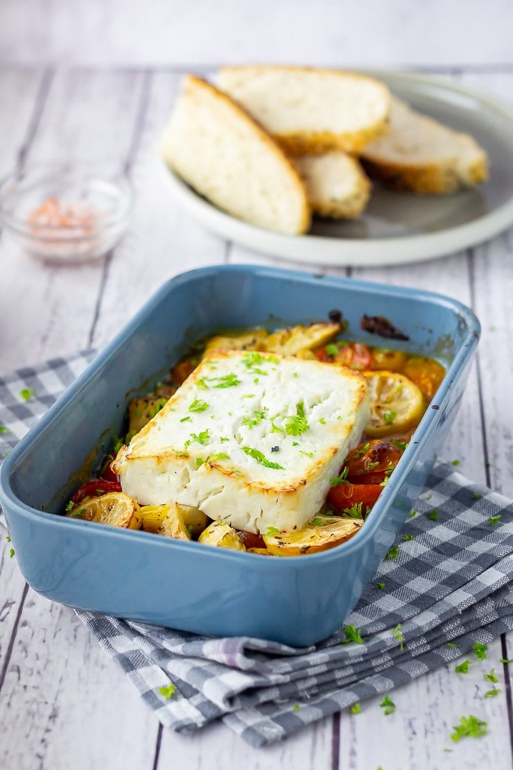 Baked feta and vegetables in a blue baking dish on a checked cloth with bread in the background