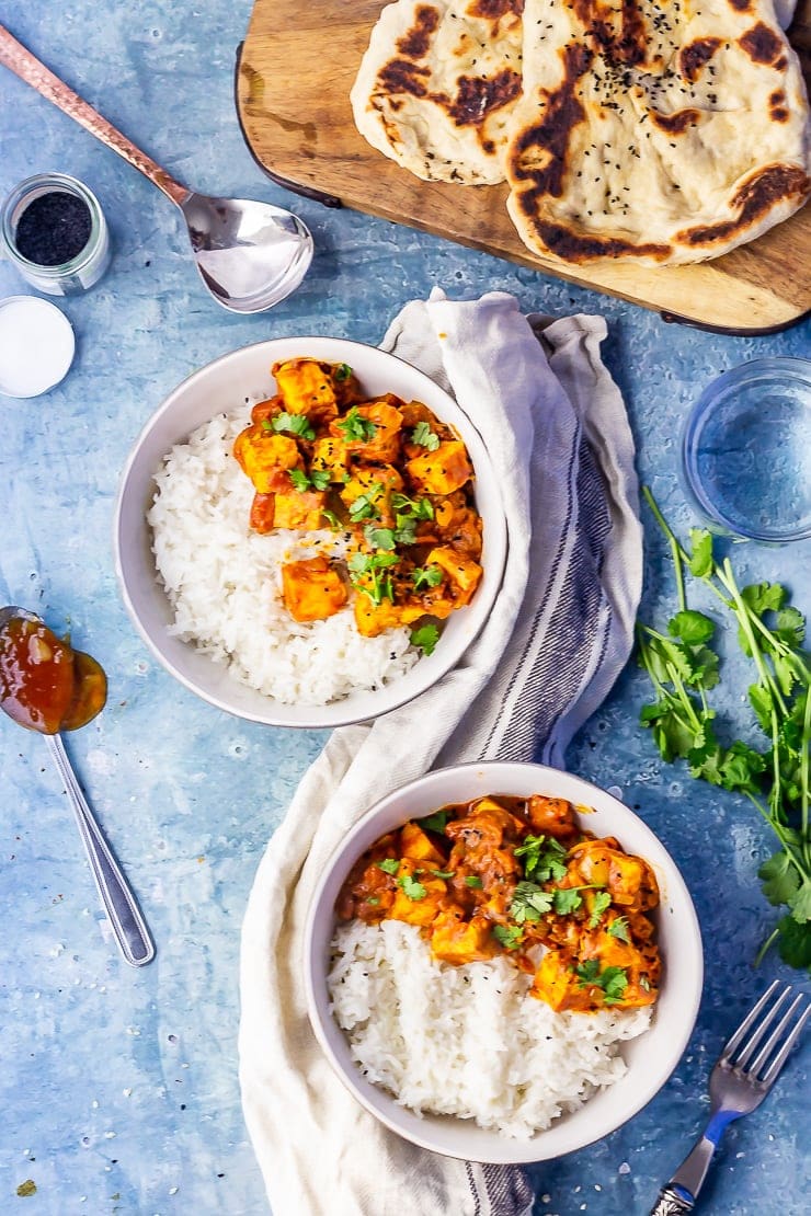 Overhead shot of paneer tikka masala on a blue background with a cloth, naan bread and spoons