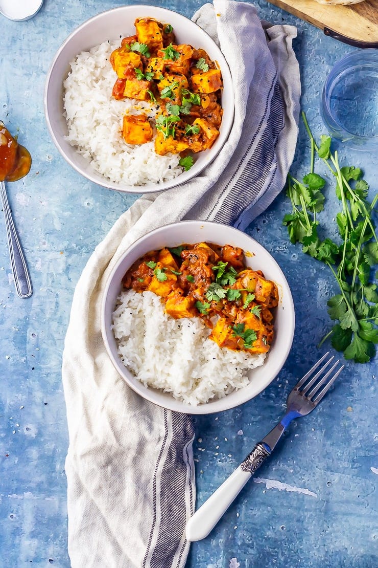 Overhead shot of two bowls of paneer tikka masala on a blue background with a cloth, fork and spoons