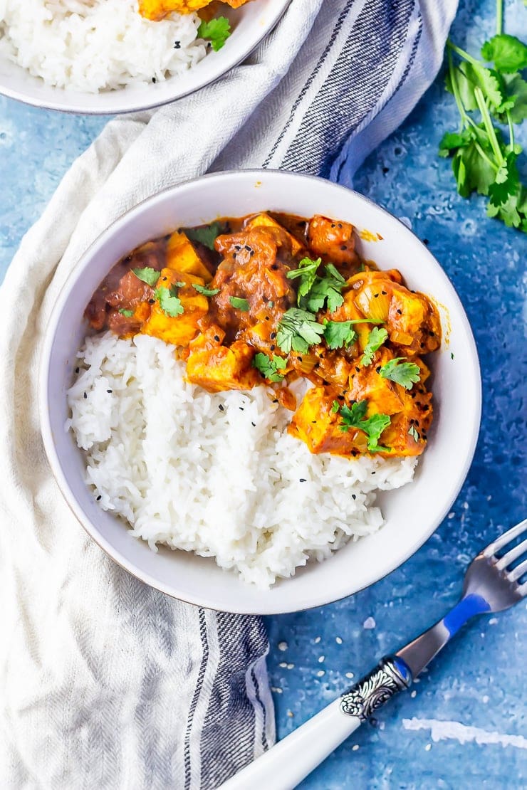 Overhead shot of a bowl of paneer tikka masala on a blue background next to a cloth with a fork